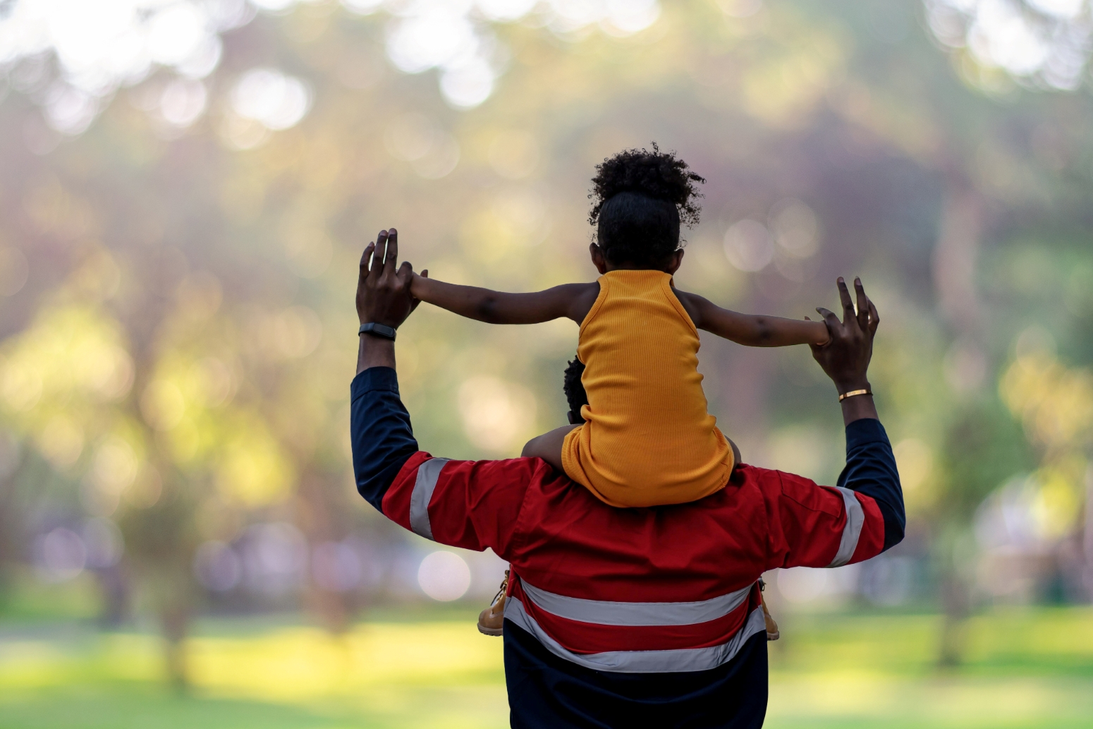 Adult with child on shoulders outside with their backs to us. They are holding hands in the air.