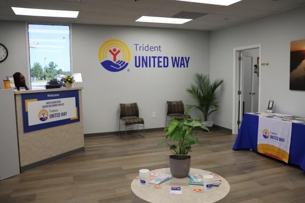 Photo of the North Charleston Center for Strong Families. There is a reception desk with a woman sitting at it on a computer, a sign on the wall with the Trident United Way logo and two chairs underneath. On another wall there is a table with pamphlets on it.