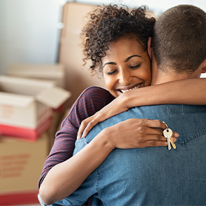 A woman is smiling and hugging a man, she holds keys in her hand and there are moving boxes in the background