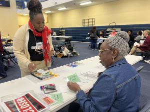 Two woman, one standing at a table and the other seated. Both facing each other, one is looking at a card in her hand