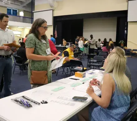 Photo of a woman standing at a folding table, holding play money and a money pouch. She is talking to two people sitting on the otherside of the folding table.