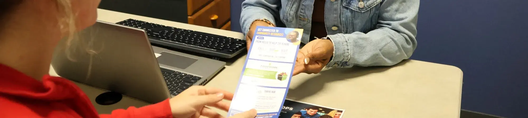 Two women sitting down, one is smiling at the other and handing her a Trident United Way rack card