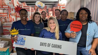 A group of 7 volunteers stand in a warehouse holding a "Trident United Way Day's of Caring" sign and are smiling at the camera.