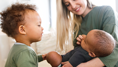 A mother sits at home with her two children. There is a baby on her lap, and a toddler looking at the baby.