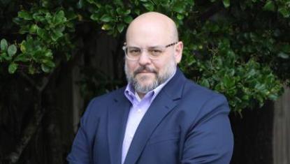 A headshot of Andrew in a blue jacket and purple shirt. He is standing in front of green trees and smiling.