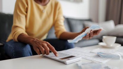 Woman in a yellow top looks at her receipt on the couch, she is punching in numbers on the calculator while other papers surround her.