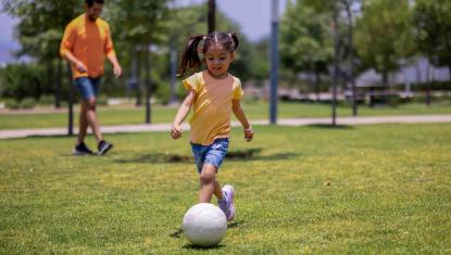 A young girl plays soccer in a yellow shirt and blue shorts. Her father is standing behind her, they are in a park playing.