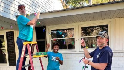 Three Days of Caring volunteers hang holiday decorations on a house and are looking at the camera smiling.