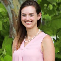Photo of Torie Brame outside in front of a tree. She is wearing a light pink tank top style blouse with a pearl necklace. Torie has straight dark brown hair. 