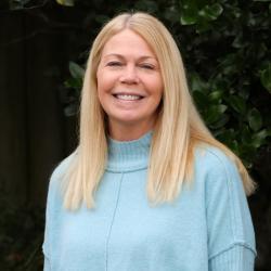 Photo of Alisha standing outside. She has blonde shoulder length hair, is wearing a blue sweater and smiling at the camera