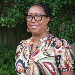 Photo of Khalilah Sumners, she is standing outside in front of the Trident United Way offices. She has on brown glasses, gold hoop earrings, and her hair is pulled back. She wears a fun patterned dress.