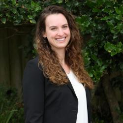 Photo of Torie outside. She has long, curly brown hair, is wearing a white top and blue blazer and is smiling at the camera.