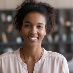 Photo of Black woman looking at camera and smiling. She is inside a book shop, and wearing a light pink top. She has short, curly hair.