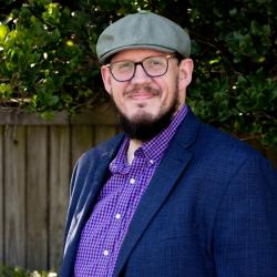 Photo of Jonathan Shettler, he is tall with facial hair, glasses, wearing a purple patterned shirt and blue jacket.