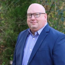 Photo of Kevin wearing a blue button up shirt, blue blazer, and glasses, smiling and looking at the camera