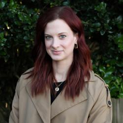 Brooke has shoulder length red hair and is wearing a black top and green jacket, standing outside smiling at the camera
