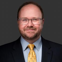 A photo of DJ standing in front of a dark grey background. DJ is smiling at the camera and wearing glasses, a black suit coat, blue collared shirt and yellow tie.