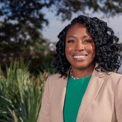 Photo of Natasha Chatman. Natasha is African American and has shoulder-length black curly hair, she is smiling and looking at the camera.  Natasha is wearing a bright green top and beige blazer, she is standing outside.