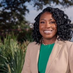 Photo of Natasha Chatman. Natasha is African American and has shoulder-length black curly hair, she is smiling and looking at the camera.  Natasha is wearing a bright green top and beige blazer, she is standing outside.